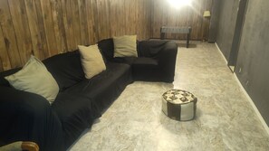 A room with a black sectional sofa, a round ottoman, a foosball table, and a wooden wall.