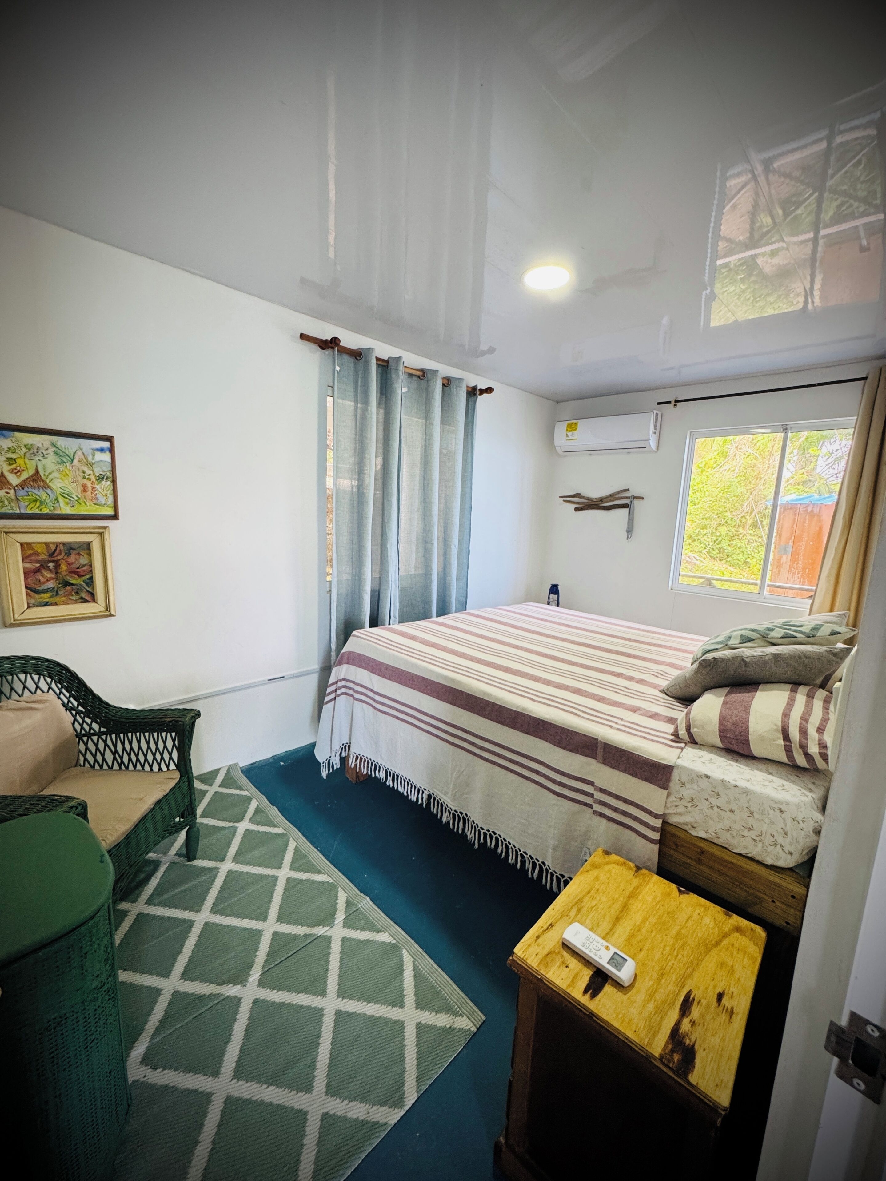 A bedroom with a bed, a chair, a small table, a window, and an air conditioning unit.