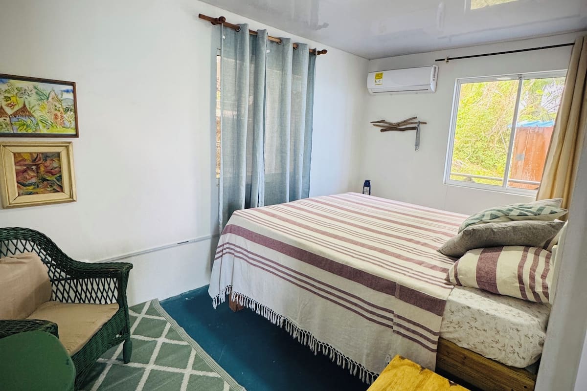 A bedroom with a bed, a chair, a small table, a window, and an air conditioning unit.