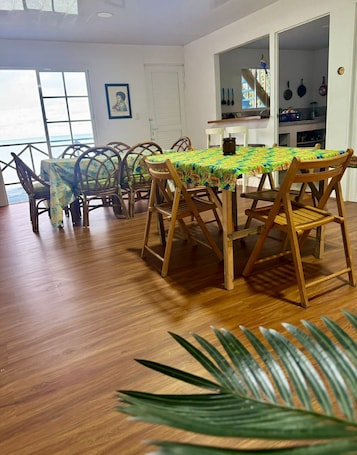 A dining area with a wooden table and chairs, a view of the ocean through large windows, and a kitchen visible through an open door.. Bay Houses Group