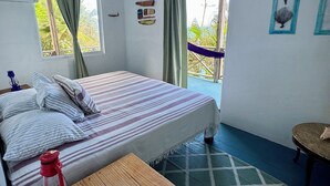 Een slaapkamer met een bed, een houten tafel, een rode waterfles, een thermometer, en een hangmat buiten het raam.
