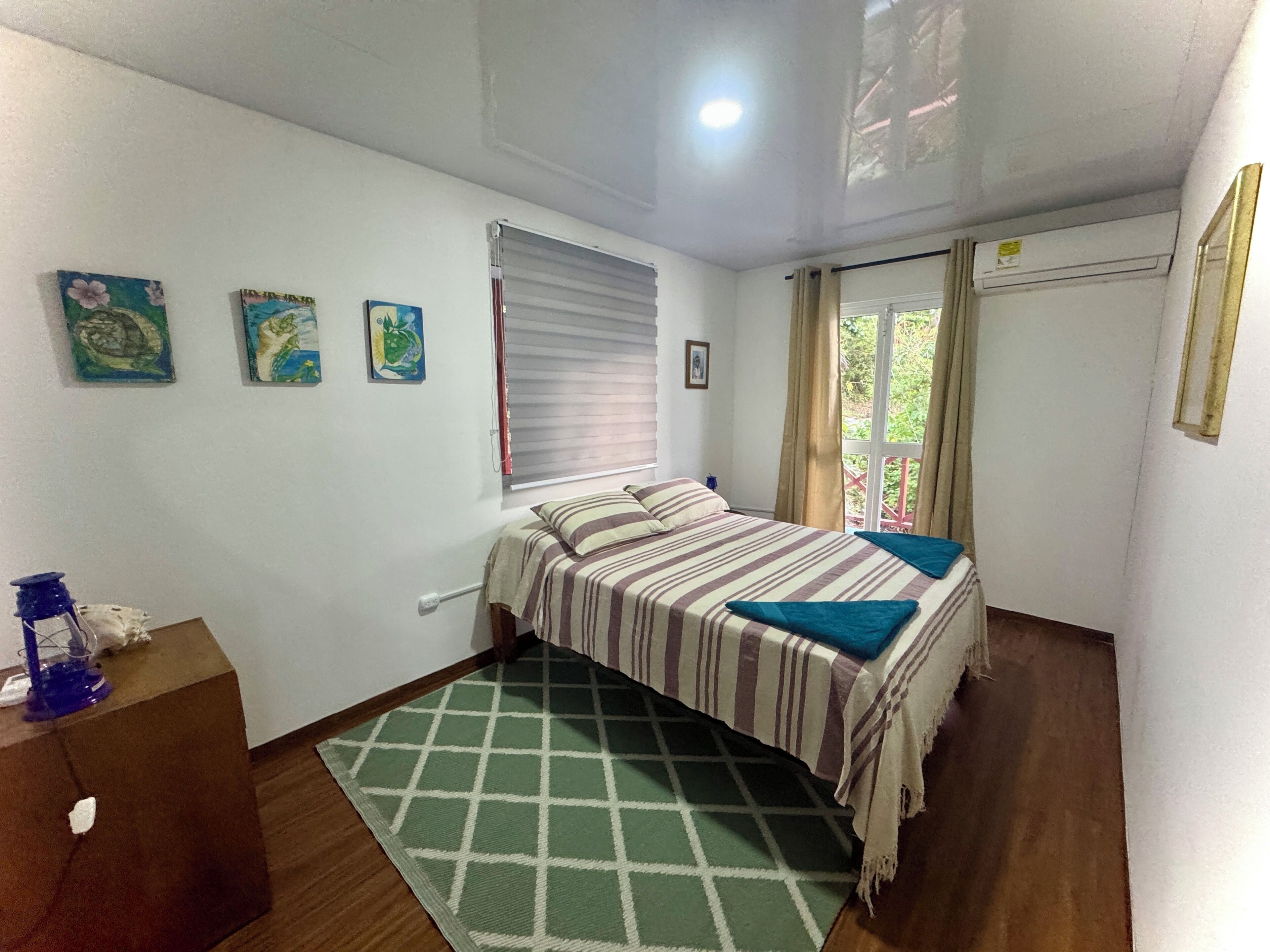 A bedroom with a bed, a window with blinds, a green patterned rug, and a wall-mounted air conditioner.