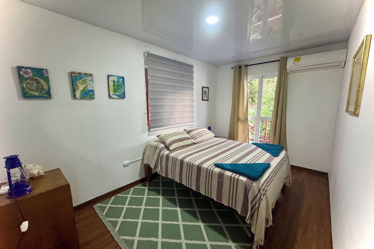 A bedroom with a bed, a window with blinds, a green patterned rug, and a wall-mounted air conditioner.