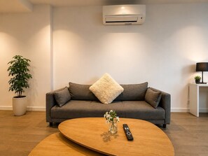 Ein modernes Wohnzimmer mit einem grauen Sofa, einem runden Couchtisch aus Holz, einer Topfpflanze und einer Wand-Klimaanlage.