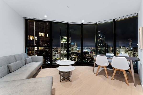 Ein modernes Wohnzimmer mit einem grauen Sofa, einem runden Couchtisch und zwei weißen Sesseln. Große Fenster bieten Blick auf die beleuchtete Stadt bei Nacht.