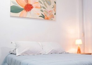 A bedroom with a bed, white pillows, a nightstand with a lamp, and a floral painting on the wall. - Spacious apartment in Las Lomas | Shopping del Sol (Asunción)