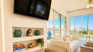 Un soggiorno moderno con una televisione a schermo piatto, un divano bianco, un poggiapiedi blu e un'ampia finestra che si affaccia su palme.