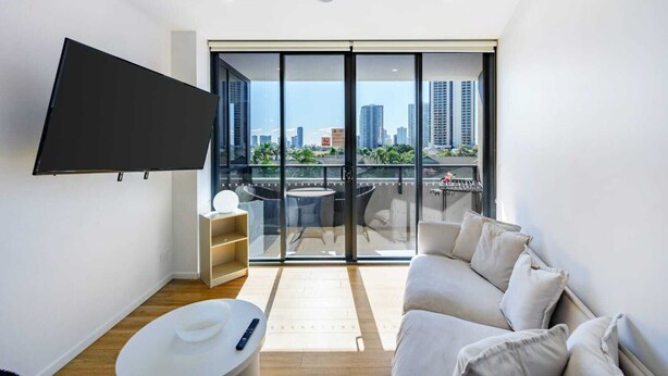 Un salon moderne doté d’un téléviseur à écran plat, d’un canapé, d’une petite table et d’un balcon offrant une vue sur la ville.