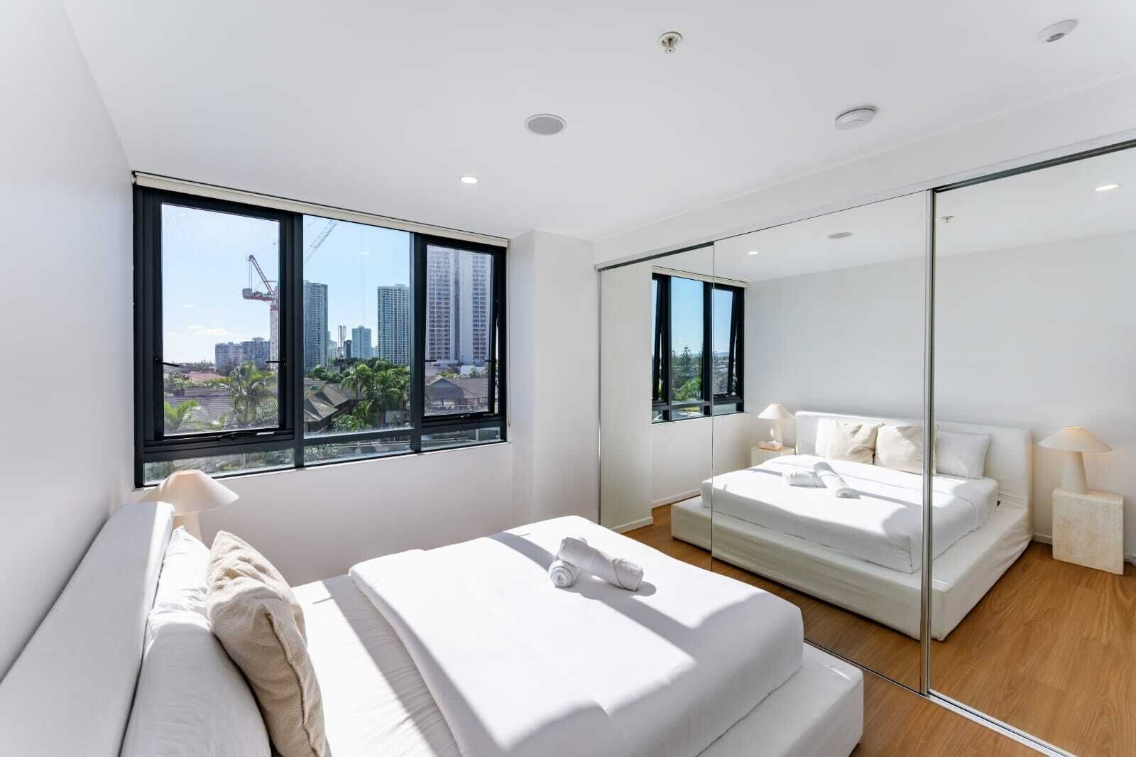 Ein modernes Schlafzimmer mit einem großen Bett, einem kleineren Bett, einem Spiegel und Blick auf die Stadt durch große Fenster.