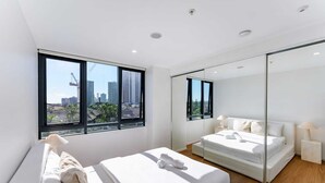 Une chambre moderne avec un grand lit, un plus petit lit, un miroir et une vue sur le paysage urbain grâce à de grandes fenêtres.