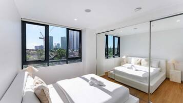 Ein modernes Schlafzimmer mit einem großen Bett, einem kleineren Bett, einem Spiegel und Blick auf die Stadt durch große Fenster.