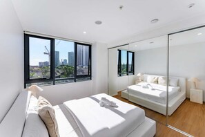 Ein modernes Schlafzimmer mit einem großen Bett, einem kleineren Bett, einem Spiegel und Blick auf die Stadt durch große Fenster.