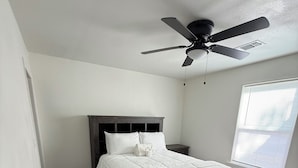 A bedroom with a bed, dark wooden floor, white walls, a ceiling fan, and a window with blinds.
