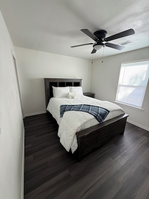 A bedroom with a bed, dark wooden floor, white walls, a ceiling fan, and a window with blinds.