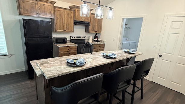 A kitchen with wooden cabinets, a black refrigerator, a marble countertop, and pendant lights.