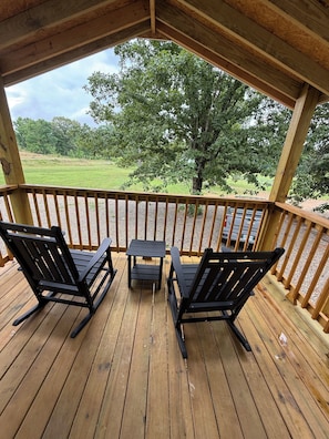 A wooden deck with two rocking chairs and a small table, overlooking a grassy area with trees.