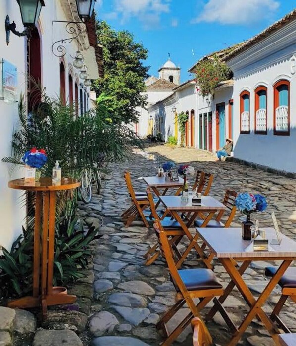 Une rue pavée avec des terrasses, des bâtiments blancs aux portes colorées et, au loin, un clocher.
