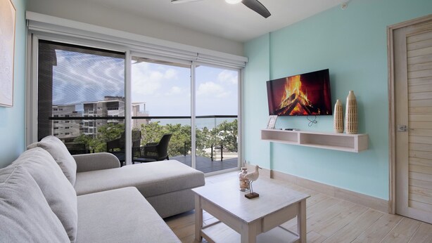 A modern living room with a sofa, a coffee table, a TV, and a balcony with a view of buildings and trees. - CASA PARAISO: Spacious 3-bedroom cozy apartment just a few meters from the ocean (Chame)