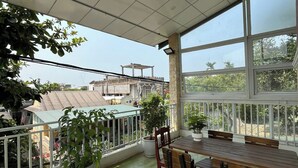 A covered outdoor area with a wooden table and chairs, potted plants, and a view of buildings and trees.