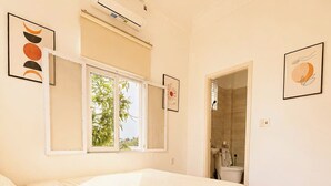 A bedroom with a bed, an air conditioning unit, a window, and a bathroom visible through an open door.
