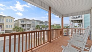 A covered porch with wooden railings, white rocking chairs, and a view of multi-story buildings. - 4 Bedroom, 4 Bathroom, Dunes Home, PET FRIENDLY (Supply)