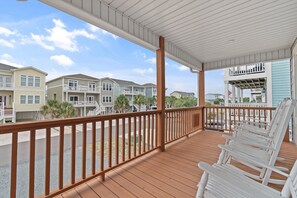 A covered porch with wooden railings, white rocking chairs, and a view of multi-story buildings. - 4 Bedroom, 4 Bathroom, Dunes Home, PET FRIENDLY (Supply)