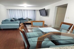 A modern living room with a glass-top dining table, blue sofas, and a flat-screen TV mounted on the wall. - Aura Bella - Prime Location! Walking Distance to Malls, Restaurants & Services. (Lima)