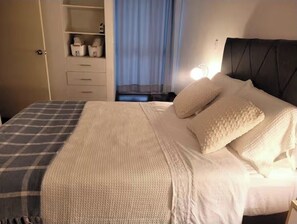 A neatly made bed with a checkered blanket, a bedside table with a lamp, and a wardrobe with storage boxes. - Aura Bella - Prime Location! Walking Distance to Malls, Restaurants & Services. (Lima)