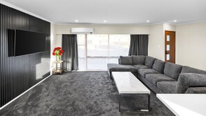 A modern living room with a sectional sofa, a flat-screen TV, a coffee table, and a large window with curtains. - Spacious 5-Bedroom Family Home Near Lake Rotorua & Rotorua CBD (Rotorua)