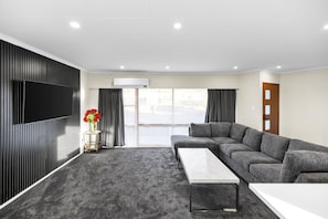 A modern living room with a sectional sofa, a flat-screen TV, a coffee table, and a large window with curtains. - Spacious 5-Bedroom Family Home Near Lake Rotorua & Rotorua CBD (Rotorua)