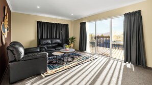 A living room with a black leather sofa, a small round table, a patterned rug, and a large sliding glass door leading to an outdoor area. - Spacious 5-Bedroom Family Home Near Lake Rotorua & Rotorua CBD (Rotorua)