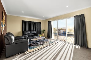 A living room with a black leather sofa, a small round table, a patterned rug, and a large sliding glass door leading to an outdoor area. - Spacious 5-Bedroom Family Home Near Lake Rotorua & Rotorua CBD (Rotorua)