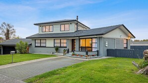 A two-story house with a garage, a front porch with potted plants, and a well-maintained lawn. - Spacious 5-Bedroom Family Home Near Lake Rotorua & Rotorua CBD (Rotorua)