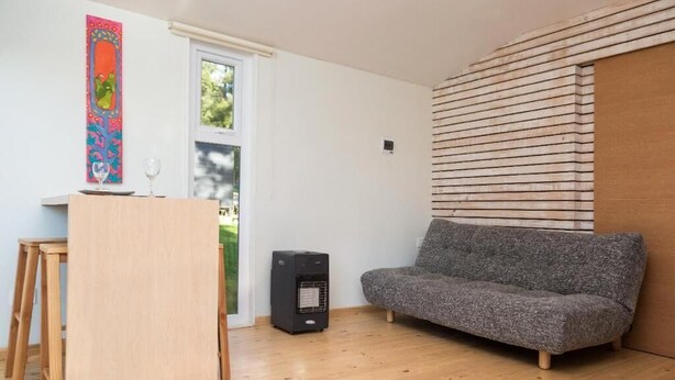 Una habitación con suelo de madera, un sofá gris, una pequeña mesa de madera, un calentador portátil y una obra de arte colorida en la pared.