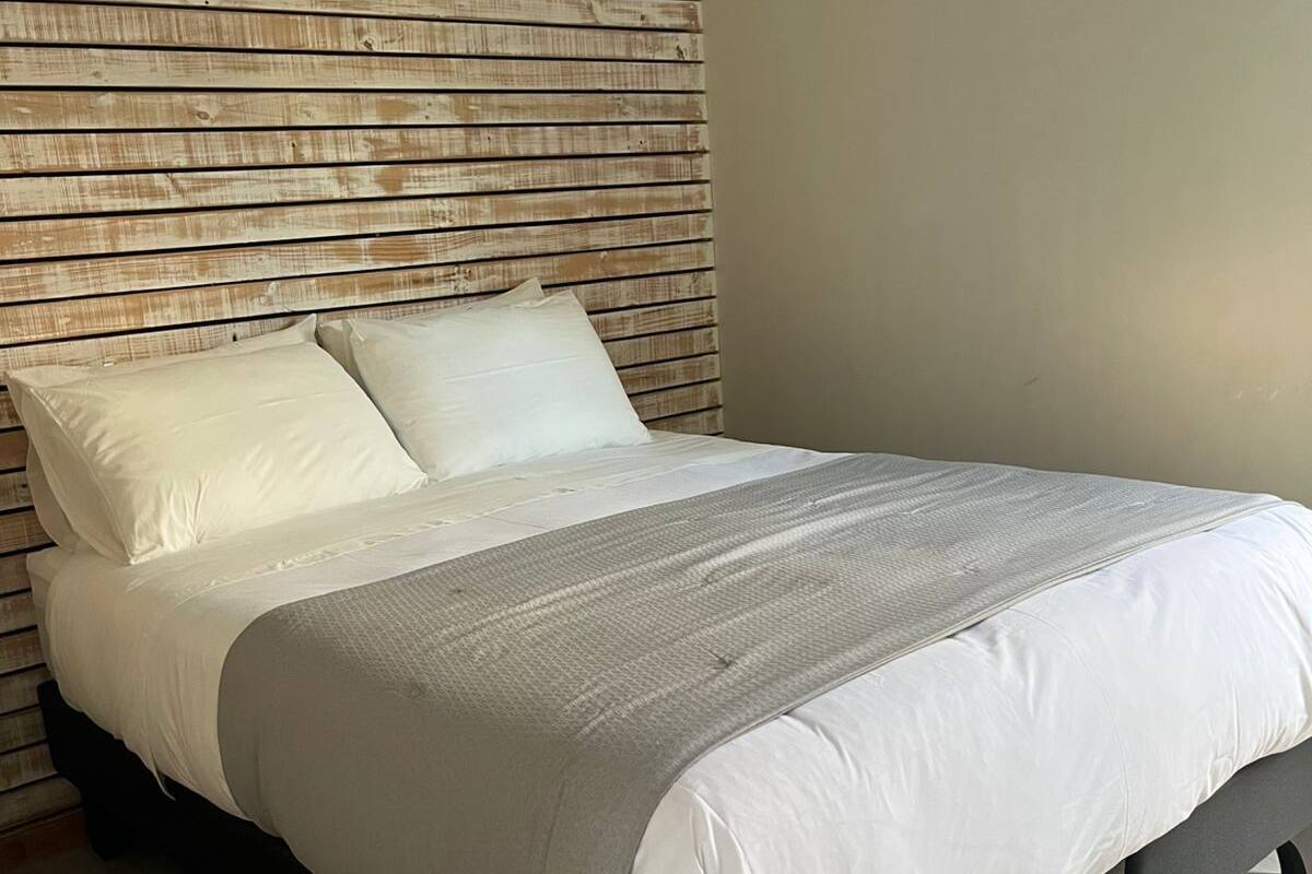 Una cama con cabecera de madera, colcha gris y almohadas blancas.