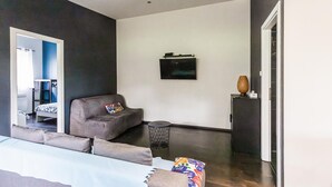 Una habitación moderna con una cama, un sillón gris, una mesita, un televisor montado en la pared y una puerta.