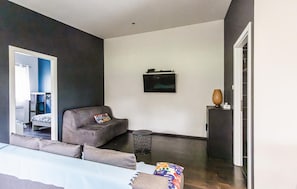Ein modernes Zimmer mit einem Bett, einem grauen Sessel, einem kleinen Tisch, einem an der Wand montierten Fernseher und einer Tür.