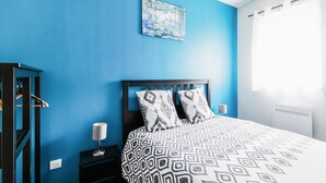 Un dormitorio con pared azul, cama con ropa de cama estampada, mesita de noche con lámpara y un cuadro sobre la cama.