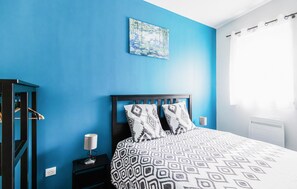 Ein Schlafzimmer mit blauer Wand, einem Bett mit gemusterten Betttextilien, einem Nachttisch mit Lampe und einem Gemälde über dem Bett.