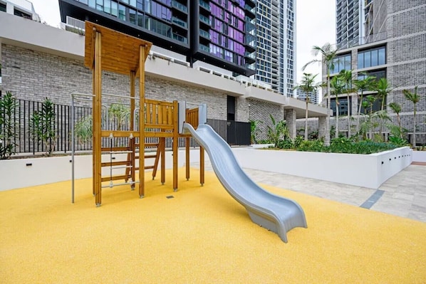 Un terrain de jeux doté d’un toboggan et d’éléments d’escalade, entouré d’une zone de jeux jaune, le tout situé devant un bâtiment moderne agrémenté de balcons.