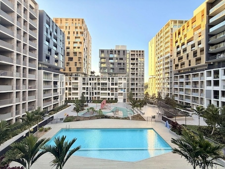 Un immeuble d’appartements moderne doté d’une piscine centrale, entouré de verdure et de palmiers.. Greenhouse Haven 1BR|Gym|Pool|Playground|Parking