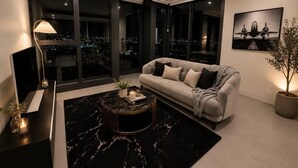 Un salon moderne avec un canapé, une table basse, un téléviseur et une grande fenêtre donnant sur la ville.