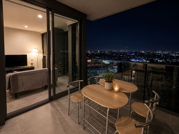 Ein Balkon mit Tisch, Stühlen, einer Couch, einem Fernseher und Blick auf die beleuchtete Stadt.