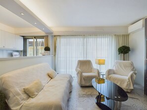 Un salon moderne comprenant un canapé, deux fauteuils, une table basse en verre et une vue sur l’extérieur à travers des rideaux légers.