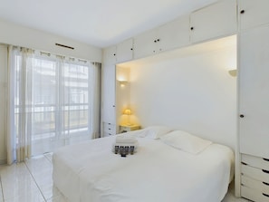 Ein Hotelzimmer mit einem großen Bett, einem Nachttisch mit Lampe und Blick durch ein Fenster mit durchsichtigen Vorhängen.