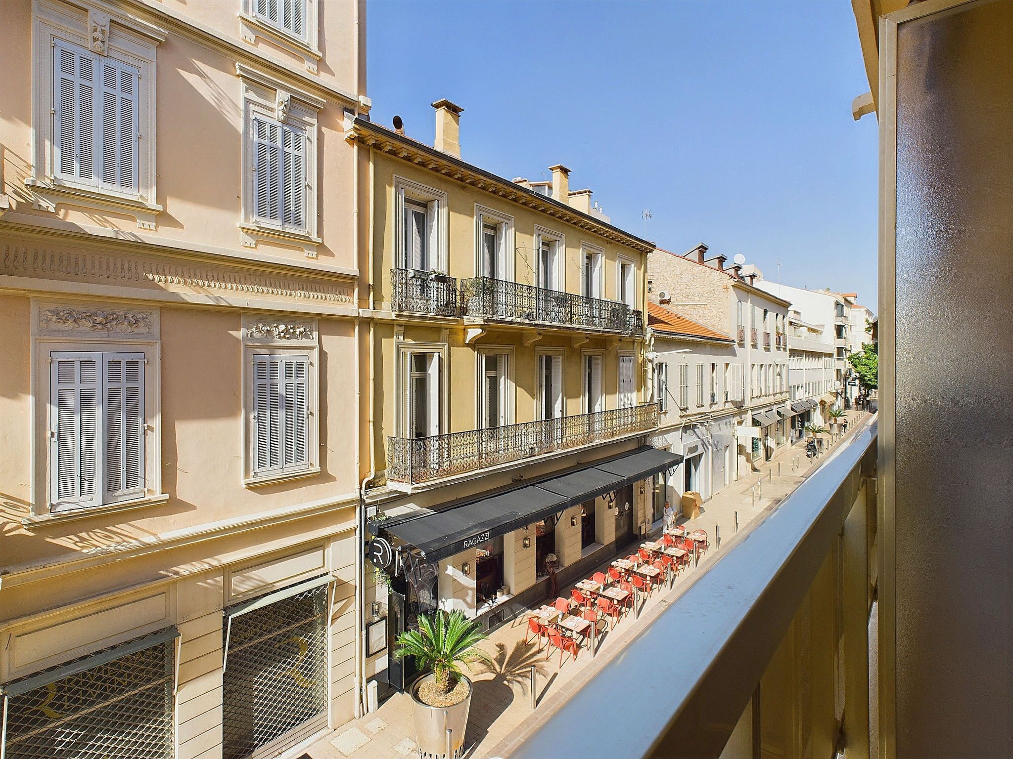 Une rue étroite bordée de bâtiments des deux côtés, un café avec des tables en terrasse et un ciel dégagé.