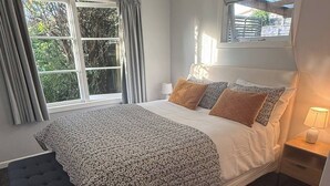 A bedroom with a large bed, a window with curtains, a bedside table with a lamp, and a view of greenery outside. - Welcome to Blue Haven in St John’s - incl pets (Whanganui)