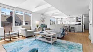 A spacious living area with a large window, a dining table, and a kitchen in the background.