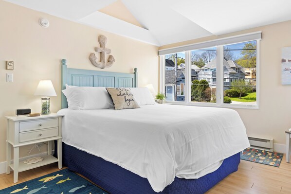 A bedroom with a bed, nightstand, lamp, and a view of houses through a window.
