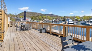 A wooden deck with tables and chairs, overlooking a marina with boats and a hilly backdrop.